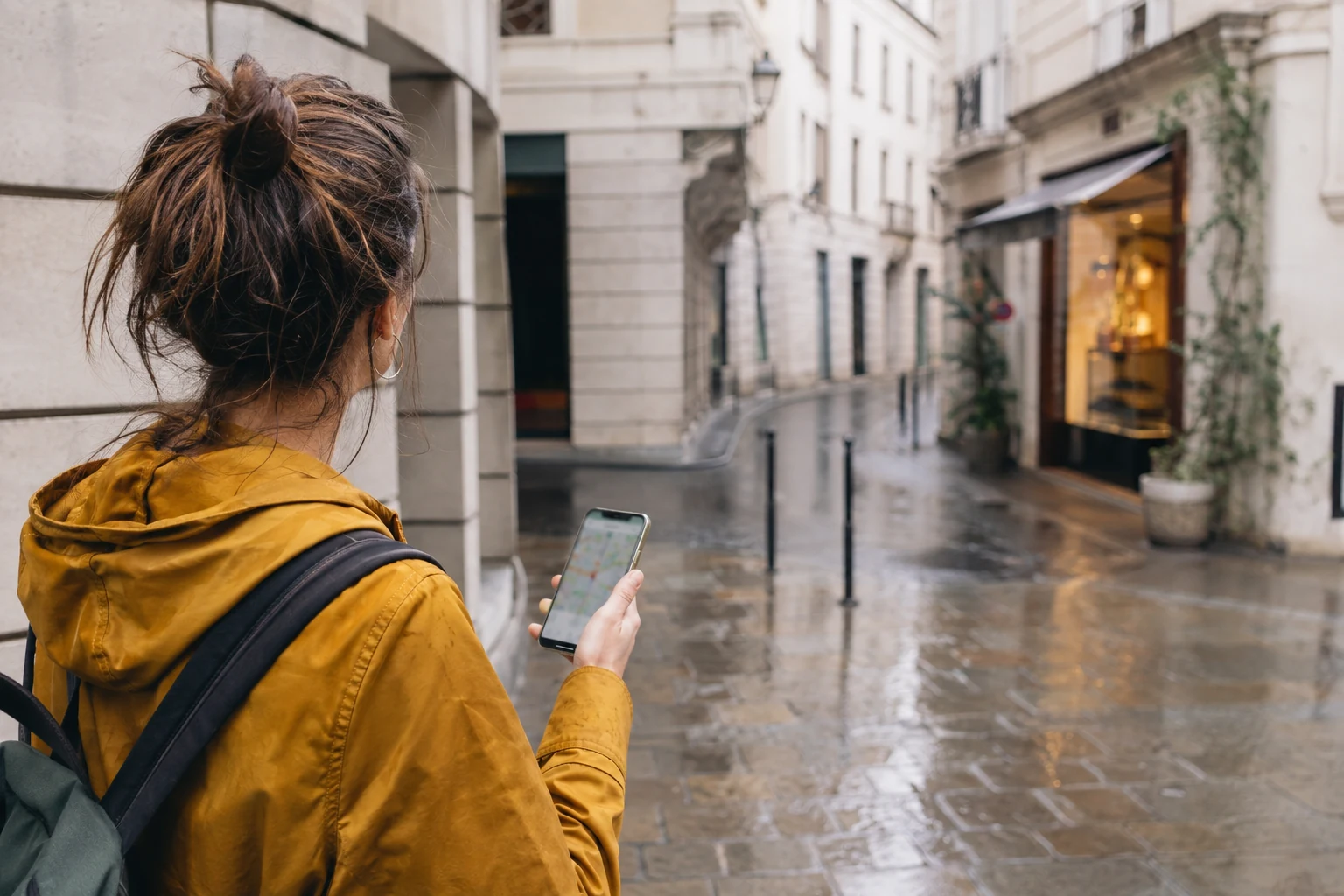 Una donna con giacca color senape consulta uno smartphone in una strada pedonale davanti alle vetrine.