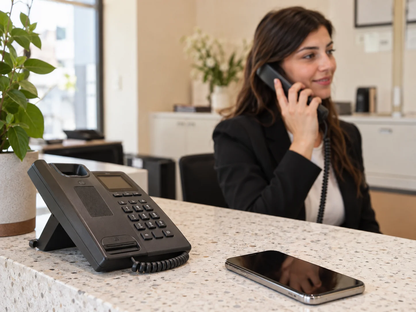 Telefono fisso e smartphone sul banco di una reception mentre una persona risponde a una chiamata