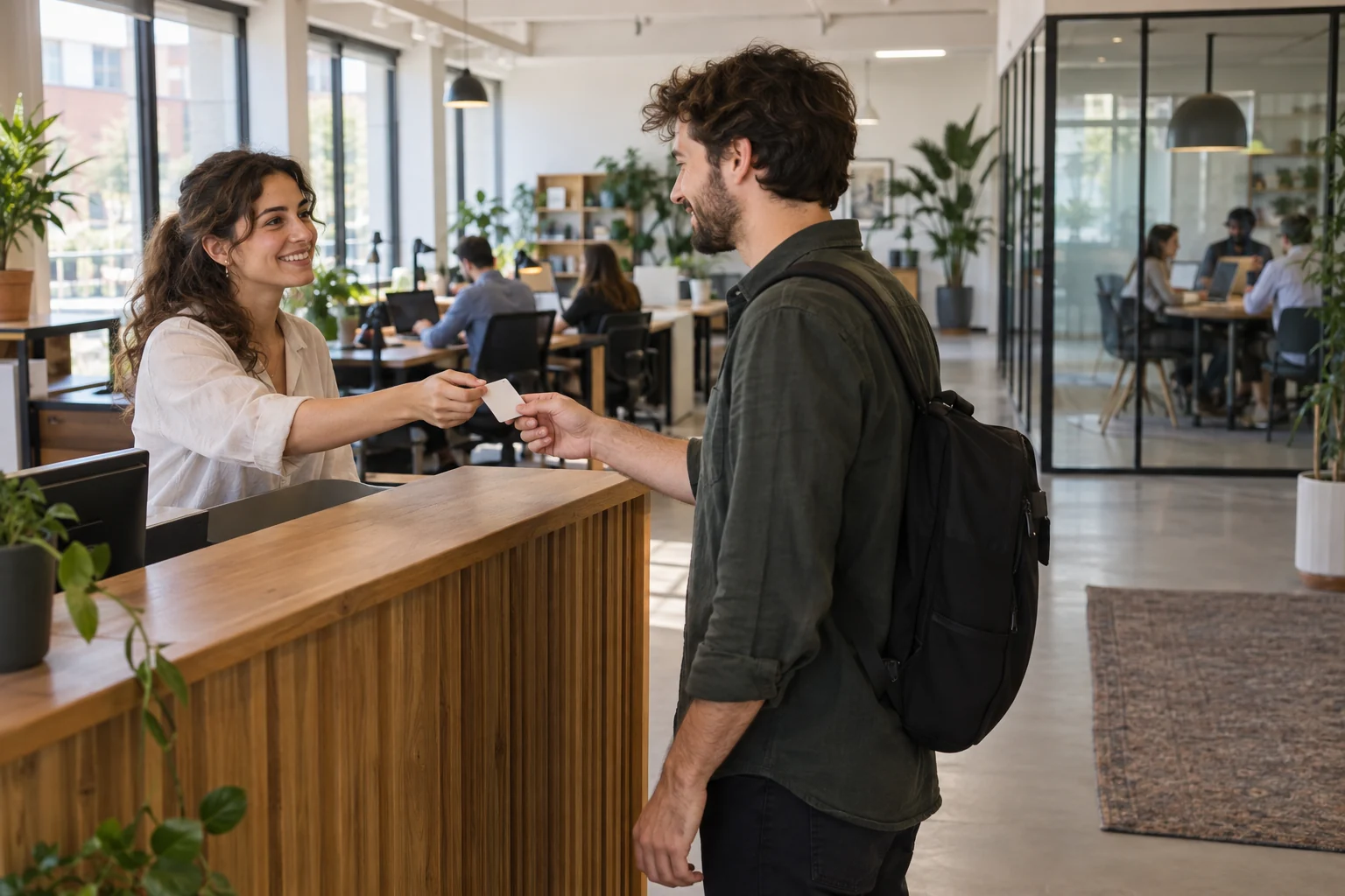 Una receptionist consegna un pass a un visitatore in un coworking con una sala riunioni visibile sullo sfondo.