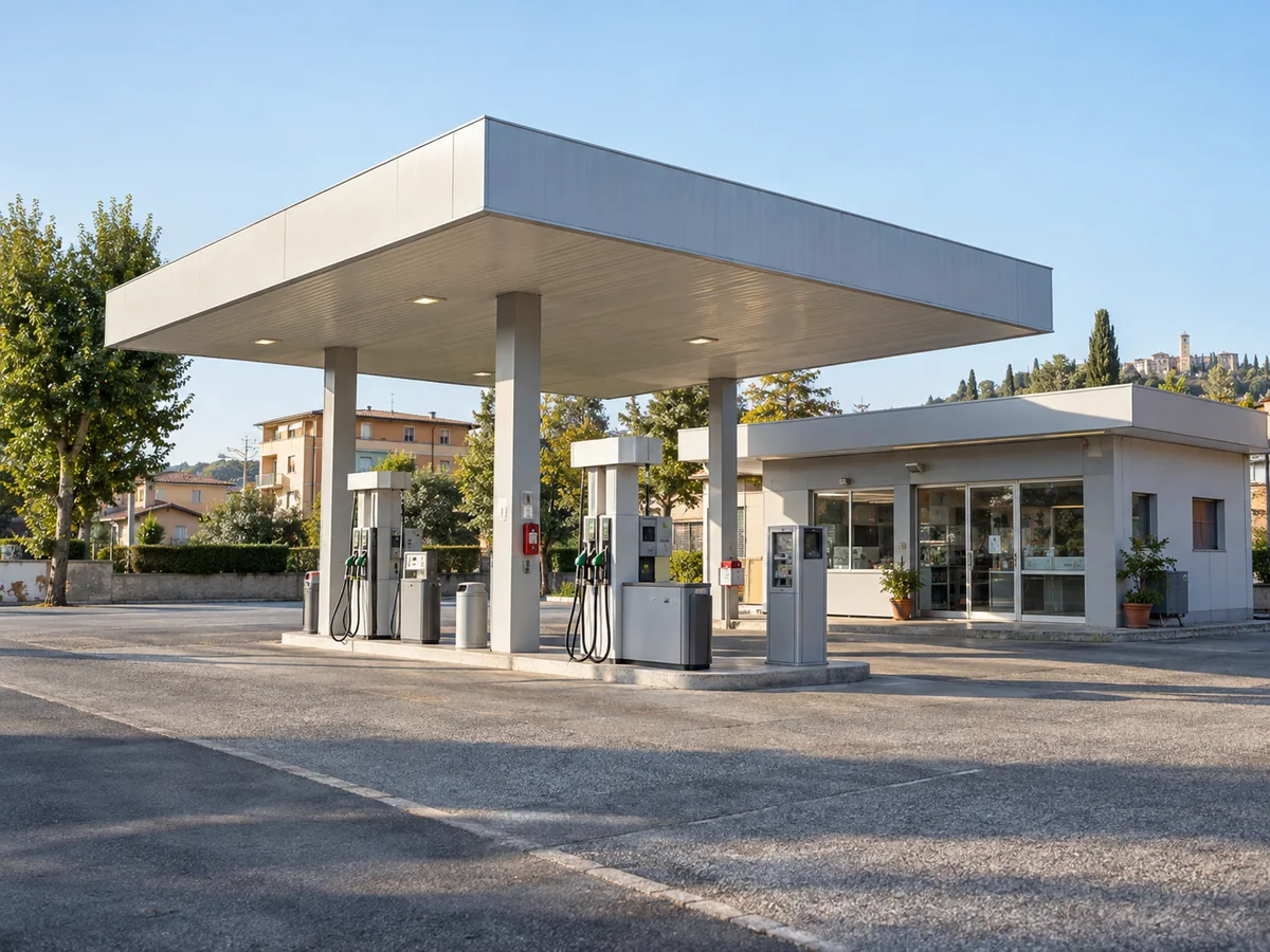 Vista diagonale di una stazione carburanti con pompe sotto la pensilina e locale di assistenza separato, nella luce del mattino.